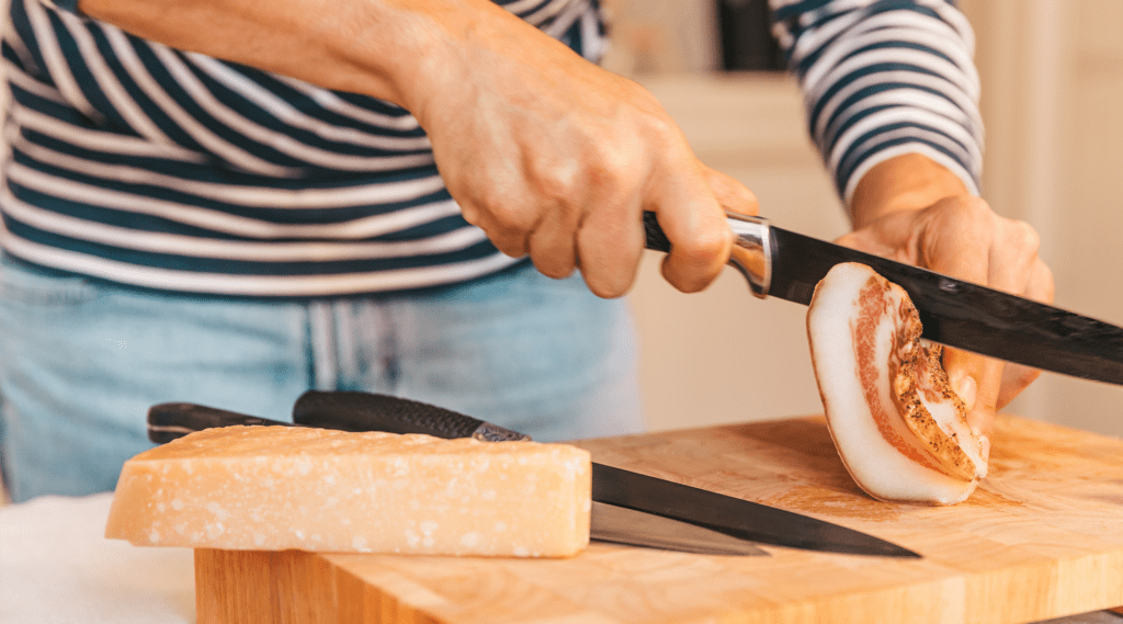 woman cutting pancetta on wooden cutting board next to hunk of cheese.