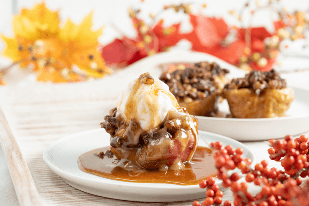 The image shows a baked nutty caramel apple on a dish in a pool of caramel sauce. The apple is topped with a melting scoop of vanilla ice cream. Two more apples are on a separate dish in the background. The image is autumnal and inviting.