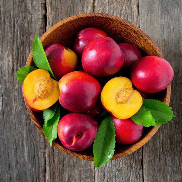 Wooden bowl of fresh whole and halved nectarines (stone fruits) on wooden table.