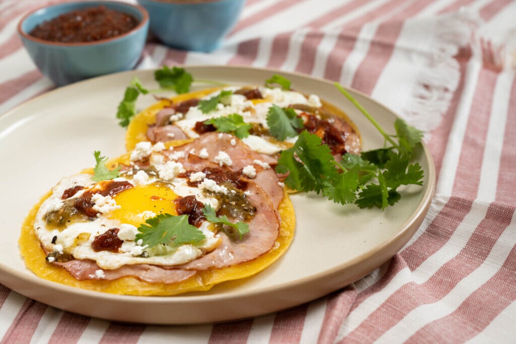 The image shows a stoneware plate with two yellow corn tortillas for huevos rancheros made with leftover ham. They have fried eggs and are garnished with cotija cheese, salsa, and cilantro.