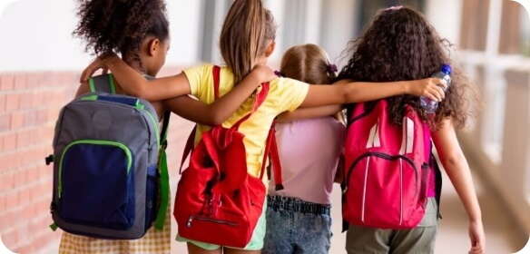 Four girls walking together with their arms around each other, wearing colourful backpacks and walking down a school hallway.