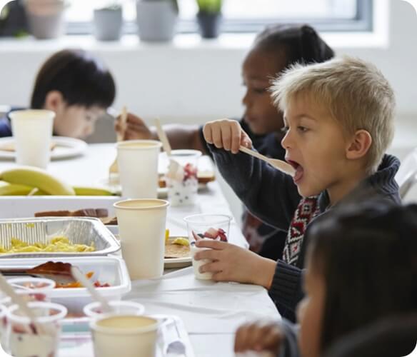 Image of kids sitting at a table and eating their lunch from containers and cups.