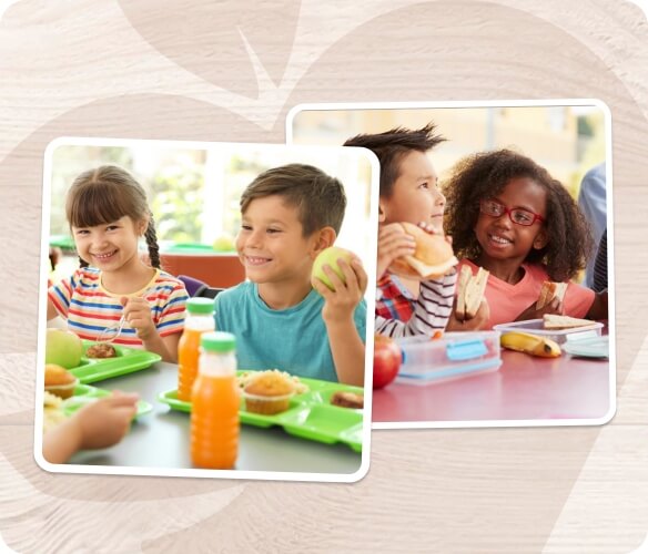 Children sitting at a table enjoying their school lunches, smiling while holding apples and sandwiches, with lunch trays and snacks in front of them.