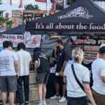 A small crowd of people walk by the event table. A few people stop to sample the free drinks.