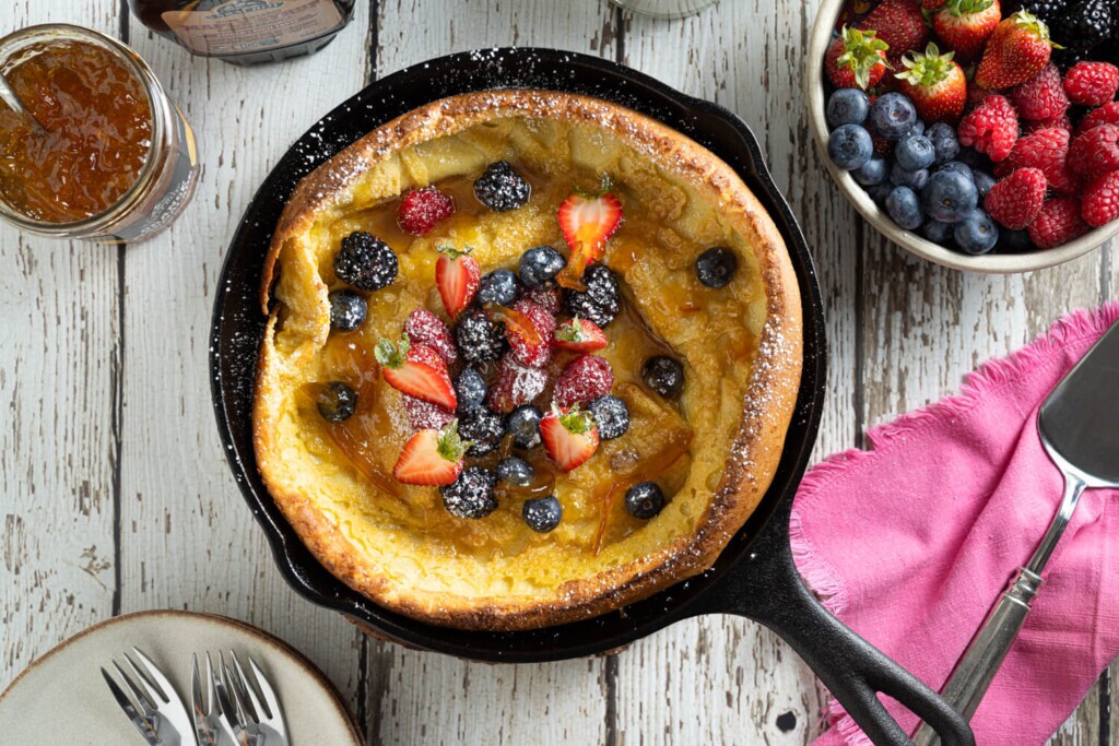 Overhead image of a cast iron skillet with a Dutch baby pancake with strawberries, blueberries, and raspberries dusted with icing sugar for brunch.