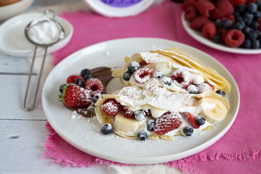 White plate on pink cloth. On the plate are banana berry crepes with raspberries and blueberries and mascarpone cream.