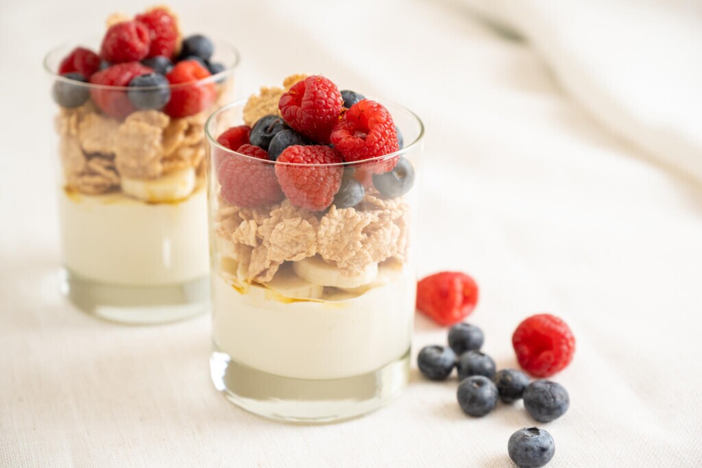 Two small glasses of brunch inspired yogurt, cereal and fruit parfaits with fresh bananas, raspberries, and blueberries.