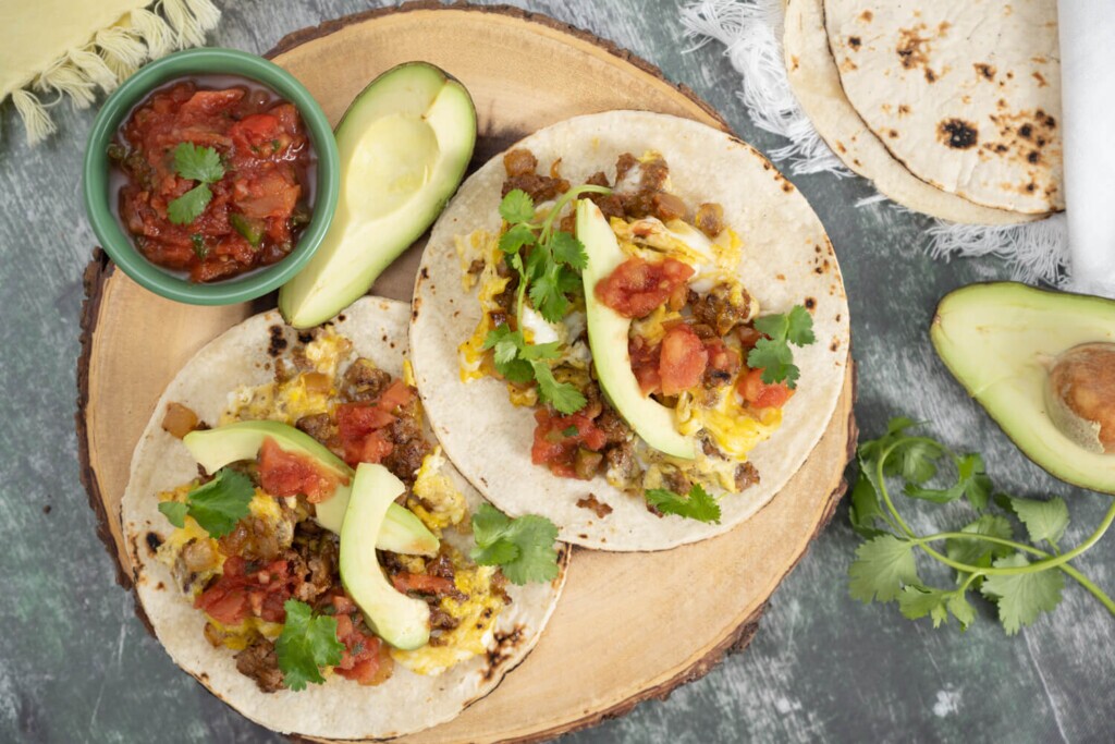 Overhead image of a cross section of log with two egg and chorizo breakfast tacos garnished with fresh avocado and salsa.