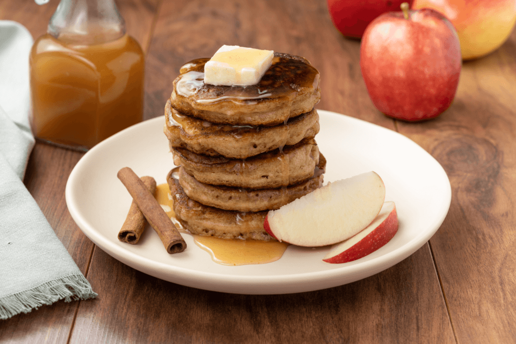 Brunch plate with stack of apple pancakes finished with a pat of butter and warm apple cider sauce, garnsihed with two crossed cinnamon sticks and two slices of apple.