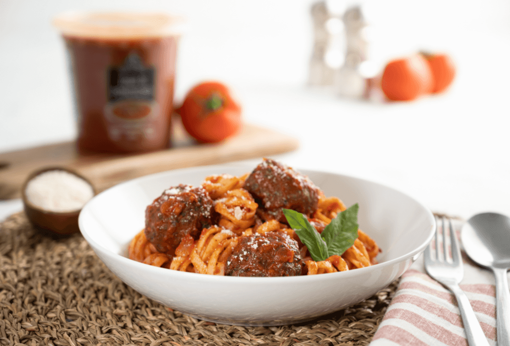 White bowl on woven placemat filled with fresh linguine pasta and meatballs with a tomato sauce and garnished with fresh basil.